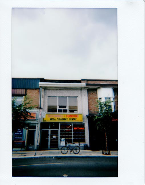 A two-story commercial building on a city street featuring a yellow sign with black lettering advertising electronics, furniture, and a mega clearance centre, with additional signage for a sectioned shop. The facade combines brickwork and light-colored panels, with a large window on the upper floor. In front of the building, a bicycle is parked on the pavement, and a black metal railing separates the sidewalk from the road. The street has two small trees planted along the footpath, and the concrete curb is visible. The sky overhead appears mostly cloudy with some light shining through, creating a soft, natural lighting environment. The scene shows typical urban surroundings, with the building's exterior indicating it could be part of a commercial area possibly managed by private disposal services such as Rubbish Clearance Chiswick, suited for alternative waste handling or rubbish clearance for local shops and flats.