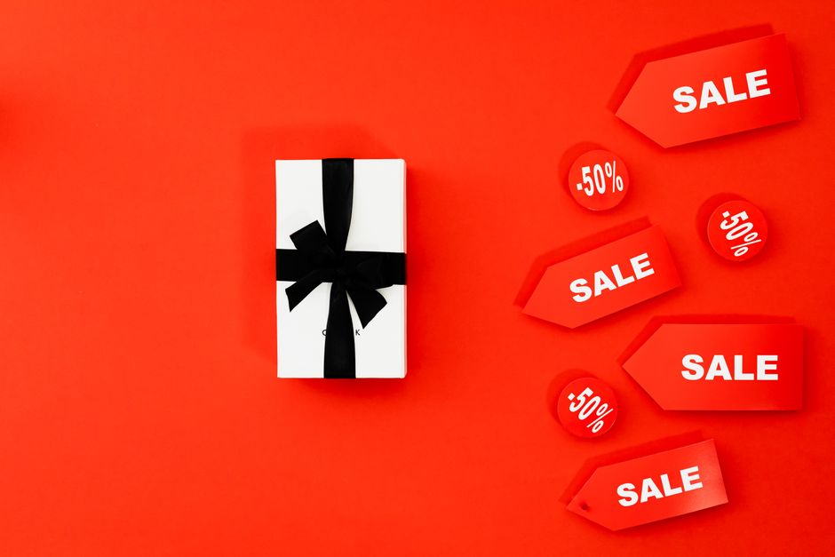 A white rectangular gift box wrapped with a smooth black ribbon tied into a bow on top, positioned centrally against a vibrant red background. Surrounding the gift are several red discount tags and circular labels with white text announcing sale percentages, such as '-50%' and '50%'. The tags are angled and layered, creating a dynamic layout with the box as the focal point. The overall setting appears to be a flat-lay composition, emphasizing promotional sale items, with no additional objects or environmental context visible. The lighting is even, highlighting the textures of the glossy tags and the matte finish of the gift box, visually connecting the scene to retail or promotional purposes, subtly aligning with themes of bargain sales and retail clearance events.