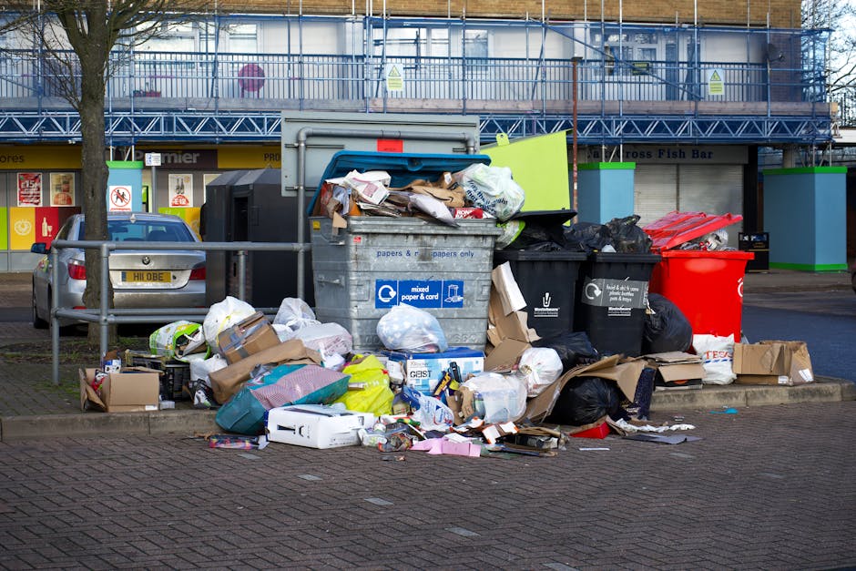 A collection of overflowing waste bins and scattered rubbish on a paved urban street corner, with a large grey mixed waste container filled to capacity positioned centrally, revealing torn cardboard boxes, plastic packaging, paper, and miscellaneous household waste spilling onto the ground. Surrounding the container are various black and red refuse bags, discarded cardboard boxes, and loose paper, creating an untidy scene. Behind the waste, a metal railing separates the rubbish area from a parking space where a silver car is parked. In the background, there is a commercial building with storefronts, including a fish bar, and a blue scaffolding structure indicating ongoing building work. The setting is outdoors during daytime, with natural light illuminating the scene, emphasizing the disorder typical of private waste accumulation and uncollected rubbish. This image highlights the importance of effective waste management practices and the potential alternatives to local authority rubbish collection, such as private rubbish removal services like Rubbish Clearance Chiswick.