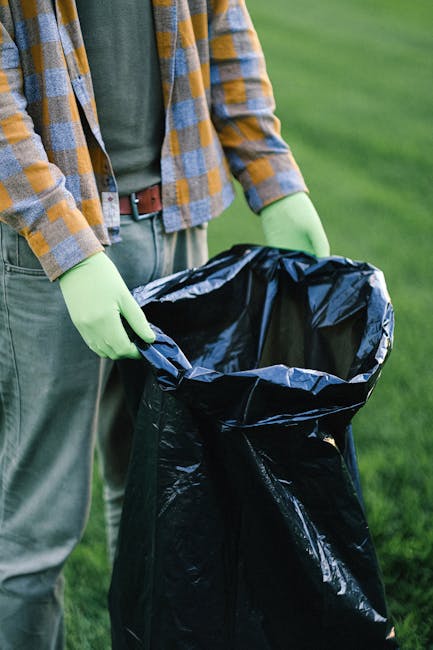 A person standing outdoors on a grassy area, holding open a large black rubbish bag lined with clear plastic. The individual is dressed in casual clothing, including a green sweater and a plaid shirt with shades of yellow, blue, and gray, and wears light green gloves suitable for waste handling. The scene shows the person's torso and arms, with the focus on the rubbish bag and their hands. In the background, there is a well-maintained lawn with a uniform green appearance, suggesting a garden or park setting. The lighting appears natural, providing clear visibility of textures, such as the glossy plastic lining of the rubbish bag and the fabric of the person's clothing. The overall scene reflects personal waste collection or garden rubbish disposal, aligning with private or non-local authority rubbish removal services like those offered by Rubbish Clearance Chiswick, supporting the context of alternative waste handling in outdoor environments.