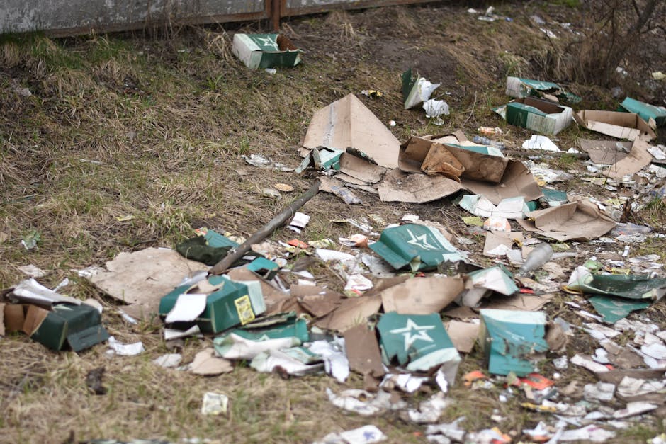 A patch of uneven ground with scattered debris including broken cardboard boxes, some with green and white star logos, and torn pieces of packaging material. The debris is spread across a grassy area near a wooden fence, with some cartons bulging and others flattened. The cardboard appears weathered, with some torn edges and creases, and a few pieces are partially collapsed. Among the rubbish, there are a few small plastic wrappers and loose paper scraps. A metal pipe or rod is visible lying across the ground, partially obscured by the debris. The surrounding environment shows sparse grass and dry patches, indicative of an outdoor space, possibly a back garden or roadside verge, where waste has been discarded. The scene reflects an example of illegal dumping or unmanaged waste collection, relevant to private rubbish clearance services such as those offered by Rubbish Clearance Chiswick, emphasizing the need for proper waste management and alternative rubbish removal options.