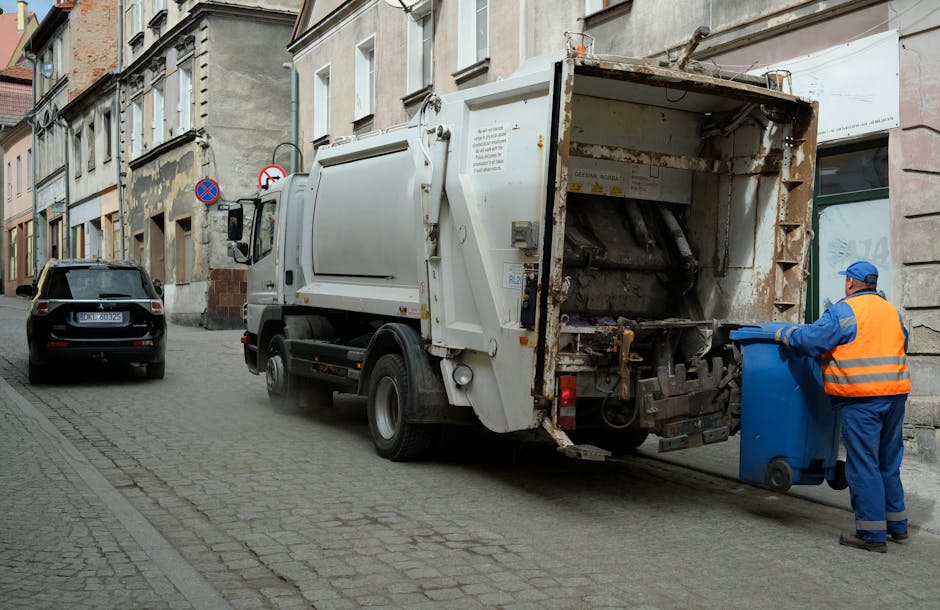 A sanitation worker wearing a blue uniform and a high-visibility orange vest is standing on a cobblestone street, emptying a blue wheeled bin into a rear-loading rubbish collection truck. The truck is white with signs of rust and wear on its metal hopper and mechanical components, positioned alongside a row of residential buildings with light-colored facades, some showing signs of aging and patchwork repairs. The street includes a parked black SUV on the left and features signage indicating parking restrictions. The scene is set during daylight with natural, diffuse lighting, highlighting the textured surfaces of the building walls, street paving, and the worker’s reflective safety gear. The image is related to private waste disposal services, illustrating an on-site rubbish collection process typical of independent waste management providers like Rubbish Clearance Chiswick, which offers alternative options to municipal rubbish collection for residential or commercial rubbish removal needs.