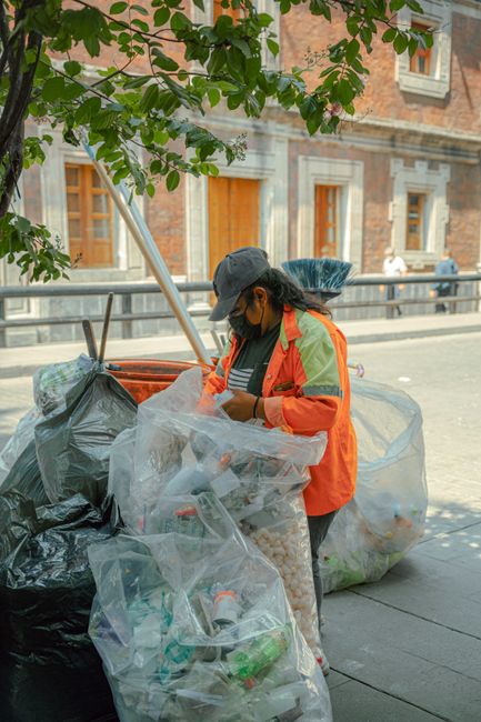 A woman wearing a dark baseball cap, black face mask, bright orange and yellow high-visibility jacket, and dark trousers is seen emptying a clear plastic bag filled with rubbish into a large black waste bag on a city sidewalk. She is standing near a tree with green leaves, which partially obscures her and provides some shade. The scene takes place in front of a historic brick building with white window frames and cream-coloured detailing around the windows. In the background, a metal railing separates the sidewalk from the street, where a few pedestrians are walking. The concrete pavement is clean and the waste disposal is organized, indicating a private rubbish collection service such as Rubbish Clearance Chiswick, often involved in on-site rubbish removal or alternative waste handling outside of standard council collection. The weather appears clear, with soft natural lighting illuminating the scene, emphasizing the straightforward act of rubbish clearance against an urban backdrop.