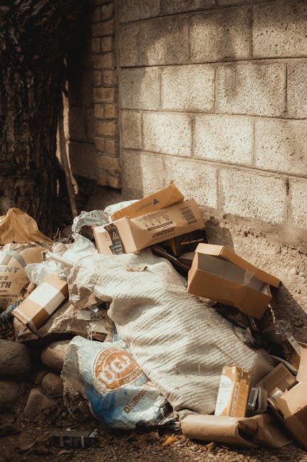 A sanitation worker wearing a blue uniform and a high-visibility orange vest is standing on a cobblestone street, emptying a blue wheeled bin into a rear-loading rubbish collection truck. The truck is white with signs of rust and wear on its metal hopper and mechanical components, positioned alongside a row of residential buildings with light-colored facades, some showing signs of aging and patchwork repairs. The street includes a parked black SUV on the left and features signage indicating parking restrictions. The scene is set during daylight with natural, diffuse lighting, highlighting the textured surfaces of the building walls, street paving, and the worker’s reflective safety gear. The image is related to private waste disposal services, illustrating an on-site rubbish collection process typical of independent waste management providers like Rubbish Clearance Chiswick, which offers alternative options to municipal rubbish collection for residential or commercial rubbish removal needs.