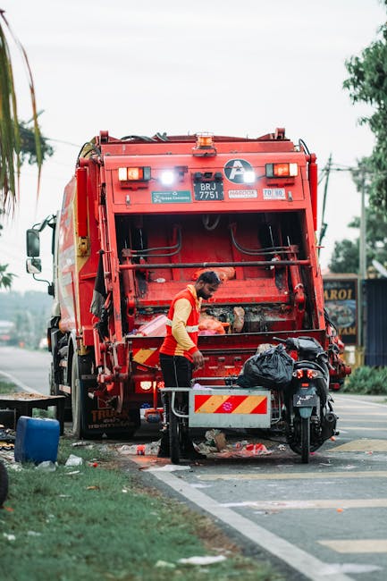 A sanitation worker wearing a yellow high-visibility vest and dark trousers is seen standing behind a large red rubbish collection truck, actively engaged in emptying the waste from a black garbage bag placed in a metal bin on the pavement. The back of the truck is open, revealing mechanized components used for waste compaction, with some debris visible underneath. The scene takes place on a suburban street, with trees and utility poles in the background, along with a building displaying signage. The pavement has a designated waste disposal area with a curb and a marked road edge. The worker appears focused, operating or inspecting the waste collection process, which is part of an independent or private rubbish removal service. The overall environment is daytime, with natural light illuminating the scene, and the setting reflects typical urban waste management activities supporting local rubbish clearance efforts.