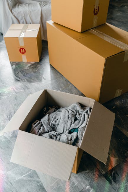 A collection of four cardboard boxes of varying sizes, situated on a mottled grey and black floor surface. The largest box, positioned prominently in the foreground, is open and contains a pile of crumpled grey fabric or clothing, with some visible blue and green elements. Two medium-sized boxes are sealed with brown packing tape and are stacked or placed nearby; one features a red and yellow circular logo with the letter 'H' visible on the top. The smallest box, located to the left, is also sealed with tape and sits partially behind the larger boxes. The background includes a section of a white sofa or cushioned furniture, suggesting an indoor environment such as a home undergoing clearance. The scene captures the process of debris or household items being prepared for disposal or collection, reflecting an aspect of independent rubbish removal services or private waste handling, as typically managed by companies like Rubbish Clearance Chiswick.